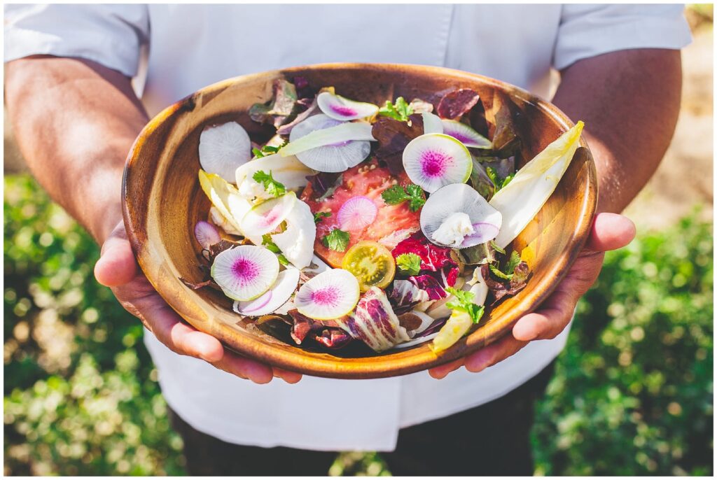 A server wearing crisp linnens holds a bowl of salad, garnished with thin slices of rainbow radishes and other fresh, local ingredients